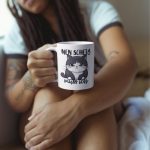 Weiße Tasse mit einer grimmig schauenden, schwarz-weißen Katze und dem handgeschriebenen Spruch „Nen Scheiß muss ich“ in Schwarz.