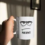 Weiße Kaffeetasse mit einem genervten Comic-Gesicht, das dicke Augenbrauen und einen schmalen Strichmund zeigt, ergänzt durch das groß geschriebene Wort „NEIN!“.