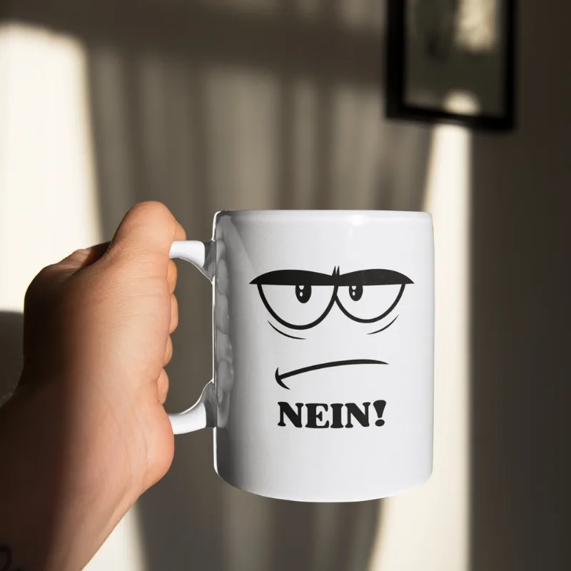 Weiße Kaffeetasse mit einem genervten Comic-Gesicht, das dicke Augenbrauen und einen schmalen Strichmund zeigt, ergänzt durch das groß geschriebene Wort „NEIN!“.